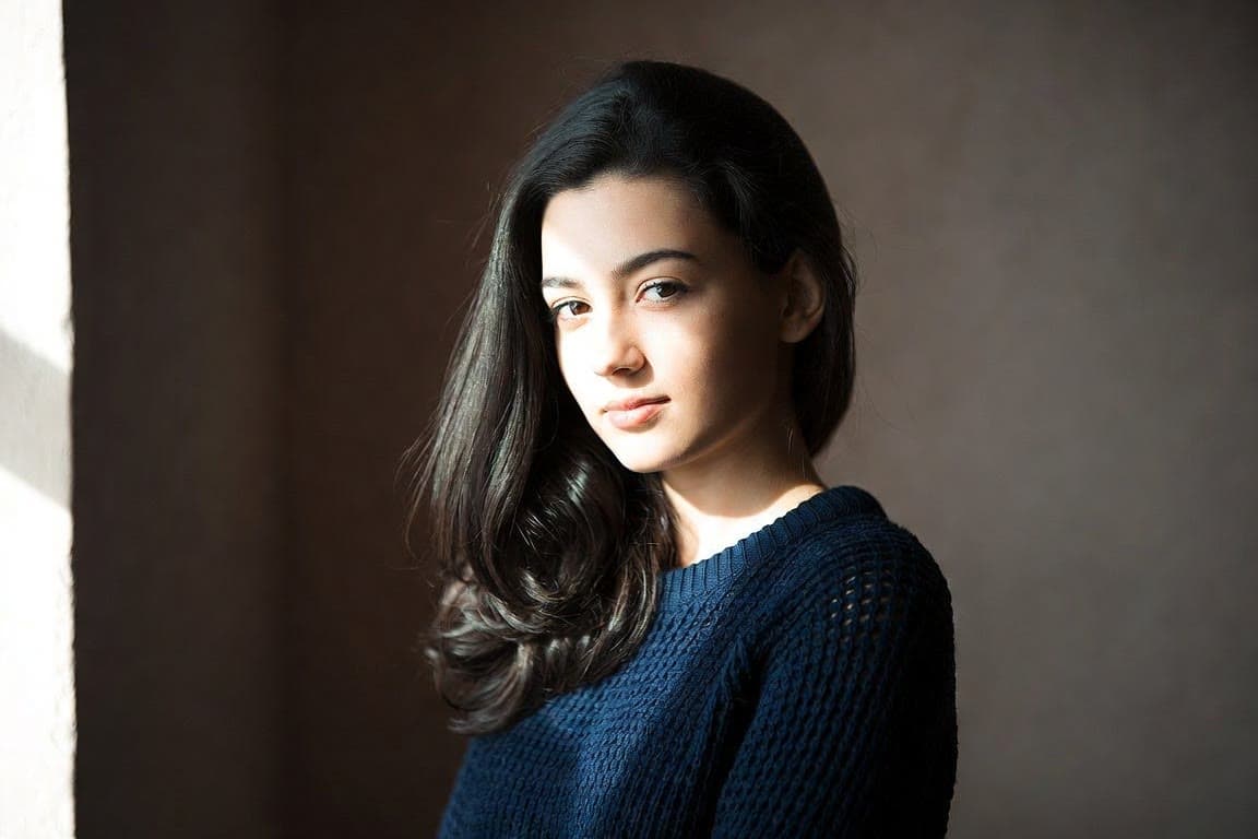 A young woman with long dark hair in a navy knit sweater, photographed against a grey backdrop.