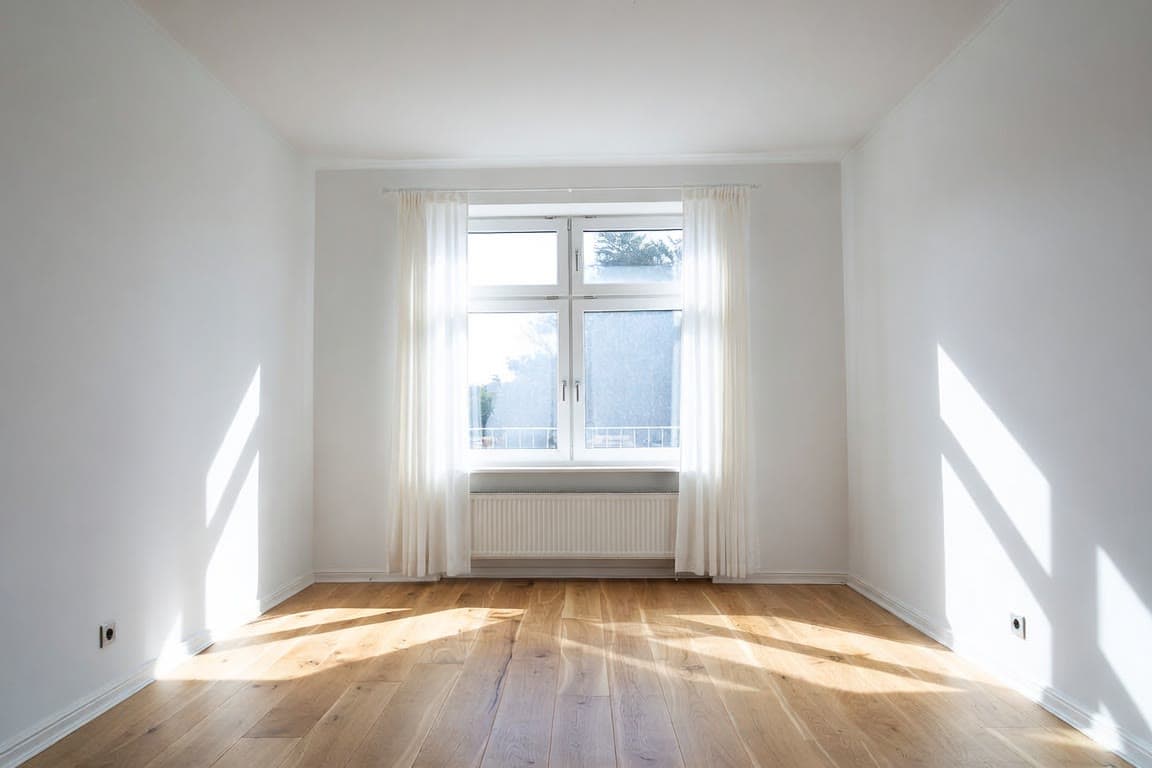 An empty living room with white walls, a wooden floor and a large window.