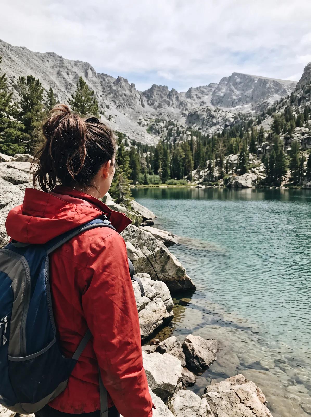 A tight portrait-shaped photo of a hiker in a red jacket at the edge of a mountain lake.