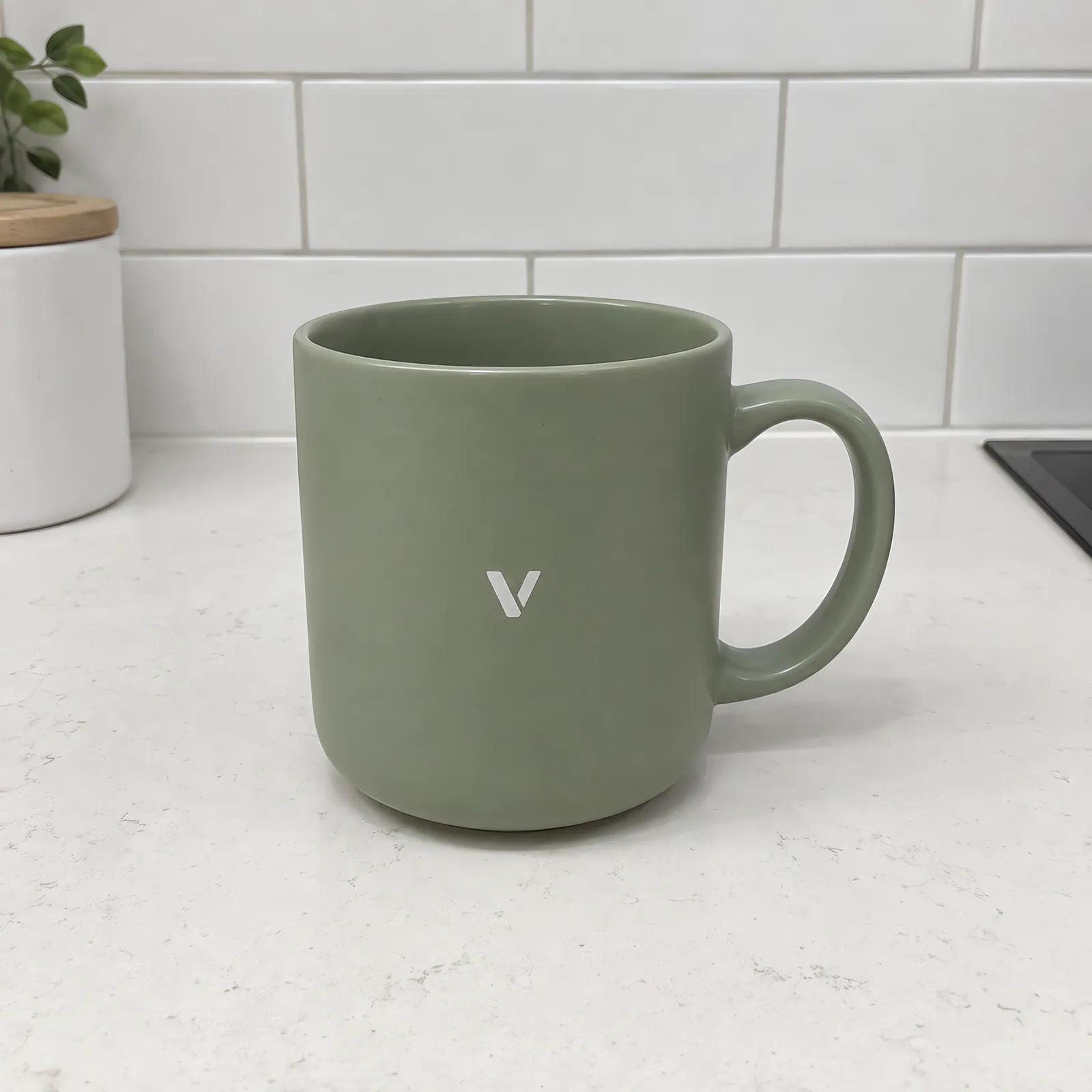 A plain photo of a sage-green mug with a small white V logo on a white kitchen counter.