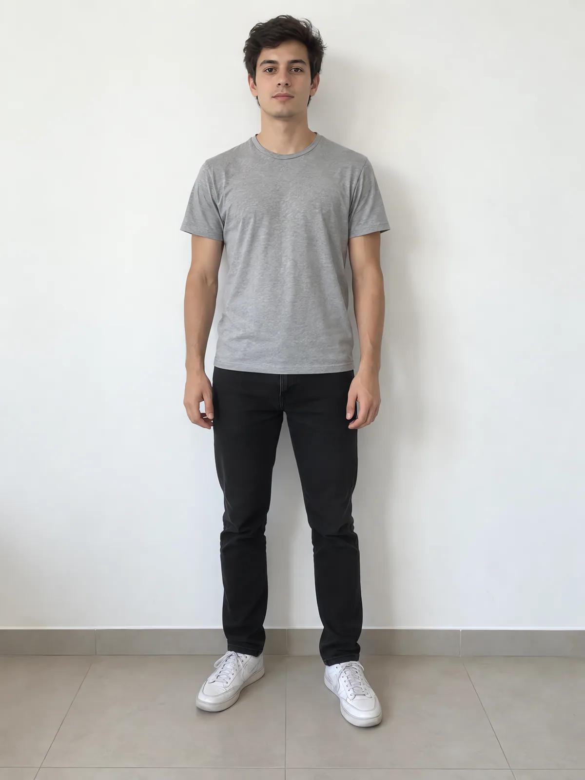 A young man in a grey t-shirt and black jeans against a white wall.